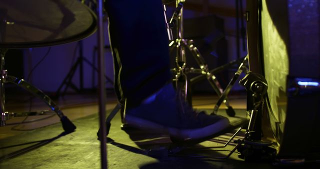 Image showcases a close-up view of a drummer's foot hitting a pedal during a live performance. The drummer's shoe is in focus while the background features the elements of a drum set under stage lighting. This image can be used to depict musical performances, instrument mastery, concert promotions, music band advertisements, or educational materials on drumming techniques.