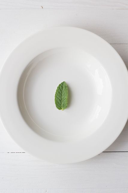 Single mint leaf placed in center of a pristine white plate, set on white wooden background. Ideal for themes of minimalistic design, culinary presentations, health food, and natural ingredients. Perfect for blogs, websites, or magazines focusing on cooking, décor, or lifestyle.