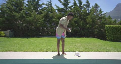 African american man applying sunscreen at pool edge on sunny summer day by backyard lawn