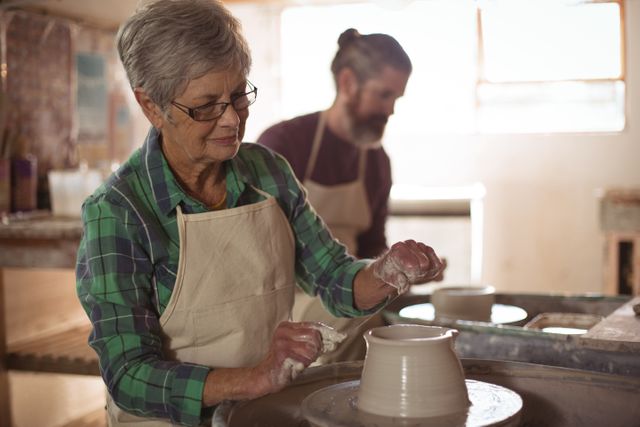 Senior woman with glasses and gray hair shaping a pot on a pottery wheel. Man working in background. Ideal for topics on crafts, pottery, creativity, senior hobbies, and art workshops.