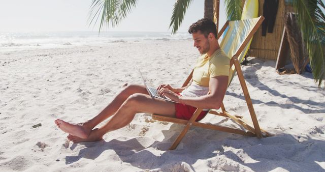 Happy biracial man sitting on deckchair and using laptop on beach. Lifestyle, realxation, nature, communication, free time and vacation.