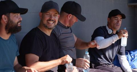 Smiling baseball teammates resting in dugout holding water bottles during game break