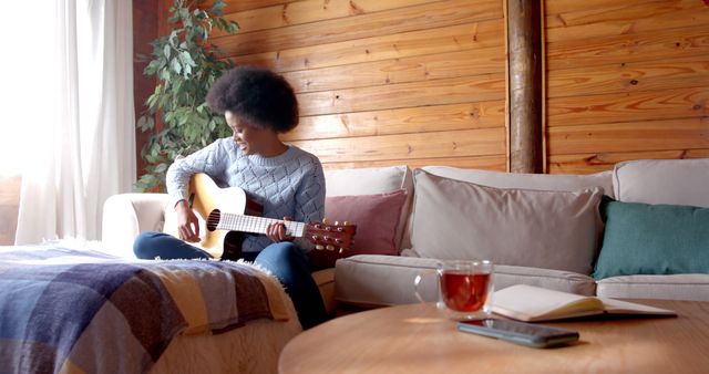Young Black woman playing acoustic guitar in a cozy living room with wooden walls and natural light coming through the window. A cup of tea and an open notebook are on a wooden coffee table, indicating relaxation and creative expression. Ideal for themes related to music, relaxing at home, leisure time, and artistic pursuits.