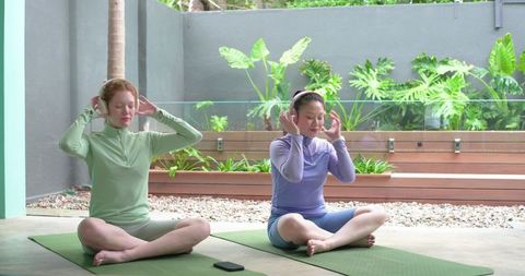Two women practicing seated meditation on tropical patio with yoga mats and headbands