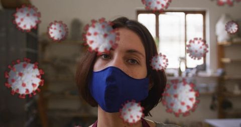 Woman wearing dark blue mask standing at pottery workbench in studio with virus graphics
