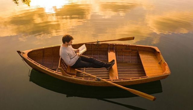 Man reading in wooden rowboat on golden lake at sunset reflecting calm solitude