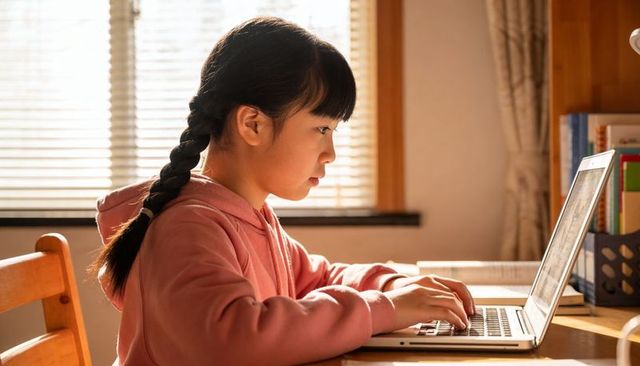 Young girl studying on laptop at home desk in warm sunlight, focused remote learning