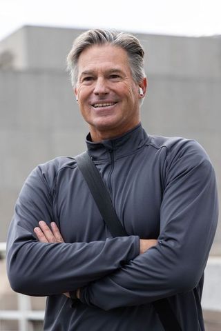 Smiling senior man wearing earbuds and athletic jacket standing on urban rooftop terrace