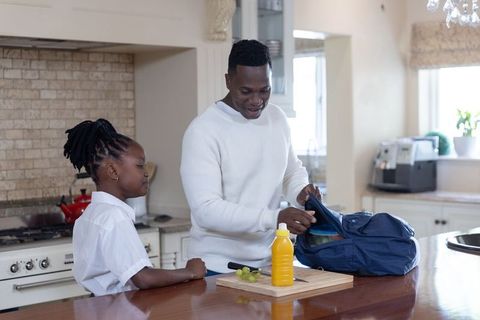 African american father packing school lunch into backpack with daughter on kitchen island
