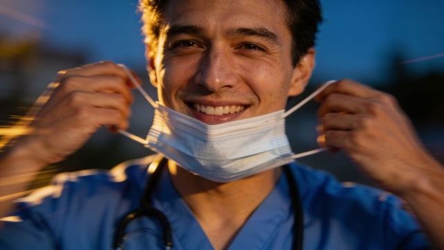 Smiling male healthcare worker lowering mask at dusk with stethoscope and blue scrubs