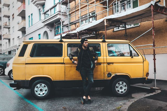 Young man wearing casual and stylish clothing leaning against a yellow vintage van in an urban parking lot. The scene features buildings and scaffolding in the background, indicating city life. Ideal for use in blog posts, travel articles, urban living concepts, hipster lifestyle, or advertisements related to road trips, exploration, and adventure.