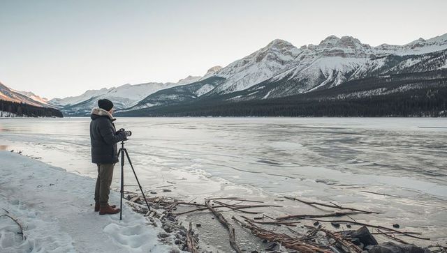 Photographer capturing frozen alpine lake and snowcapped peaks with tripod sunrise