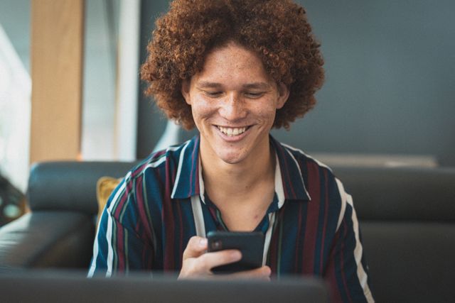 Young biracial businessman with curly hair sitting on a sofa in a business lounge, smiling while using a smartphone. Ideal for use in business, technology, and lifestyle contexts, showcasing modern work environments and professional communication.
