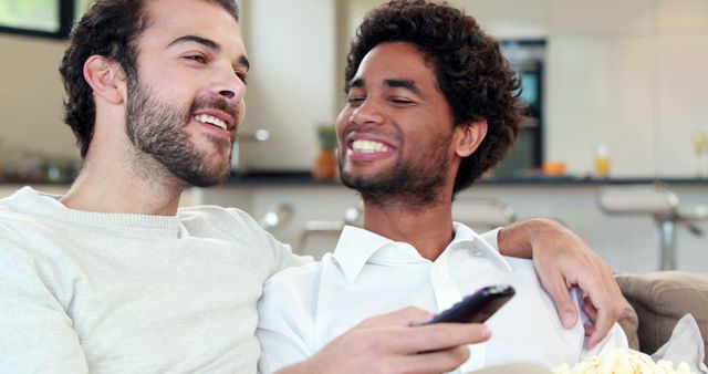 This image shows two men sitting closely on a couch in a living room, enjoying time together while watching television. One man holds a remote control while both are smiling and interacting warmly. Ideal for use in articles or advertisements focused on relationships, leisure time, home lifestyle, and inclusive branding.