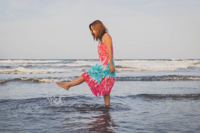 Young woman is enjoying a carefree moment at the beach, splashing water while wearing a colorful tie-dye dress. Ideal for promoting summer travel, vacation destinations, beach holidays, and relaxed lifestyle. Perfect for use in travel brochures, summer advertising campaigns, social media posts, and lifestyle blogs.