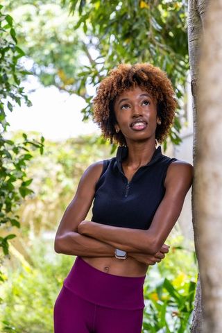 Sunlit african american woman leaning on tree, looking up, wearing crop top and leggings