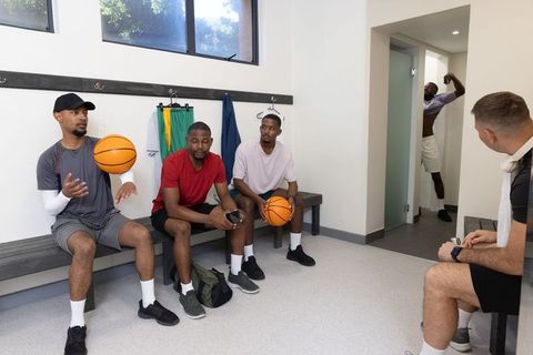 Diverse basketball teammates sitting on bench tossing basketballs and checking phone in locker room