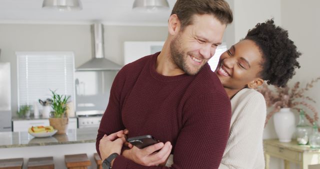 Image of happy diverse couple hugging and using smartphone in kitchen. Love, relationship and spending quality time together at home.