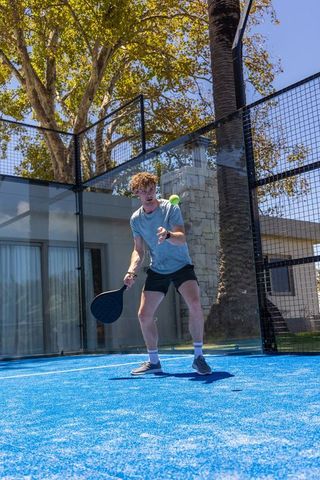 Young man playing padel on vibrant blue court with glass walls under sunlit palm trees