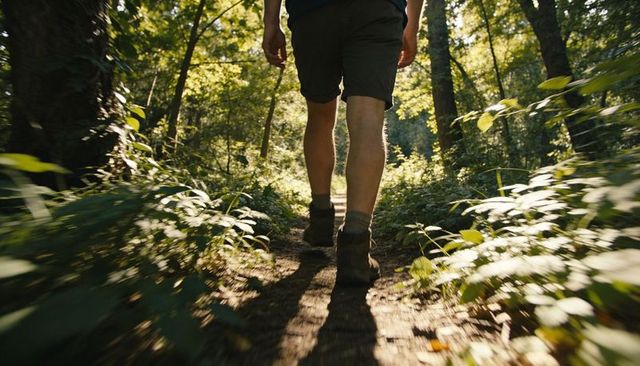Hiker walking sunlit woodland trail wearing boots and khaki shorts, dappled light