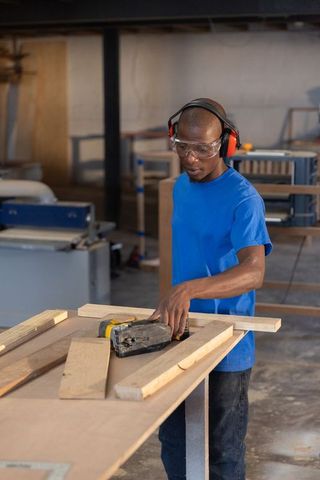 African american carpenter operating electric planer on wood planks in industrial workshop