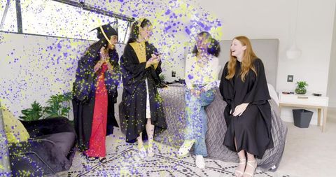 Celebrating graduates in gowns and mortarboards in chic apartment bedroom with confetti