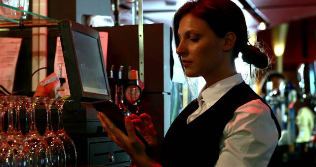 Waitress in uniform using a tablet to take an order in a dimly lit bar with red lighting. Various drink glasses and bar equipment can be seen in the background. Suitable for websites about the hospitality industry, bar or night club promotions, restaurant services, technology in service industry, and workplace environments.