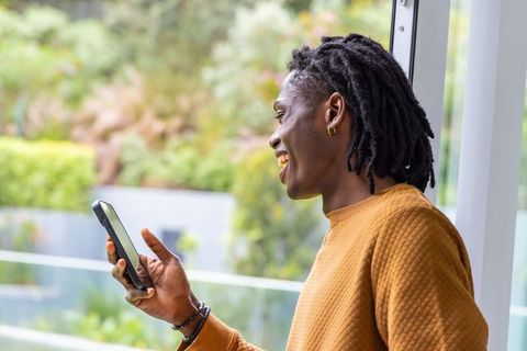 African American man smiling while using smartphone on balcony in mustard quilted sweater
