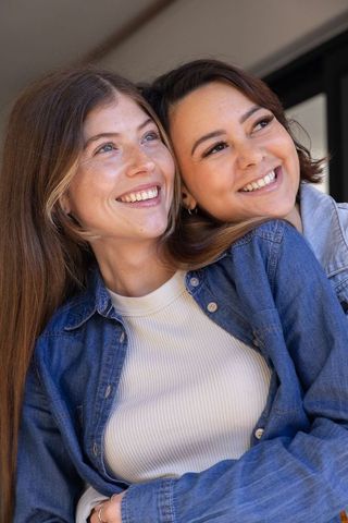 Smiling diverse female friends hugging and leaning near window in denim jackets under soft daylight