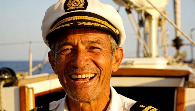 Smiling senior captain wearing peaked cap and epaulettes on sunlit sailboat deck