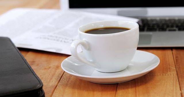 A cup of coffee sits on a desk near an open laptop and a newspaper, with copy space. It suggests a morning routine or a break during work hours, with a focus on staying informed and energized.