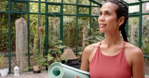 Woman Holding Mint Yoga Mat Gazing Upward in Sunlit Greenhouse for Mindful Yoga