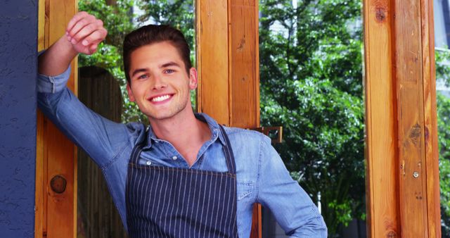 Smiling Young Cafe Worker in Denim Shirt Leaning Against Wooden Frame - Download Free Stock Images Pikwizard.com