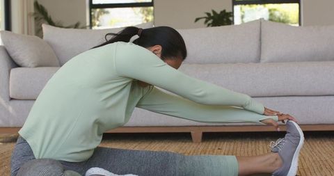 Asian woman performing seated forward stretch on woven rug for home fitness, flexibility