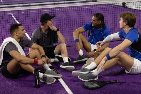 Diverse male pickleball teammates sitting at net on purple court, chatting and relaxing
