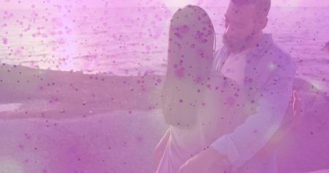 Romantic couple embracing at ocean edge during pink sunset with speckle bokeh overlay