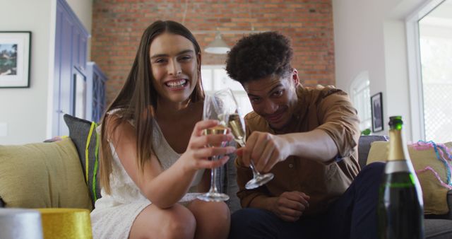 Happy biracial couple drinking champagne at home making celebration laptop image call and laughing. celebration, festivity and communication technology at home.