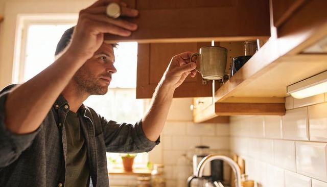 Man reaching for ceramic mug in sunlit kitchen cabinet, morning routine, cozy home
