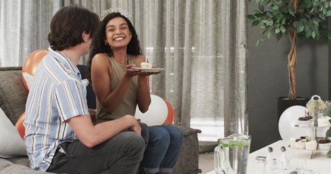 Smiling woman wearing tiara holding cupcake celebrating birthday with partner on sofa