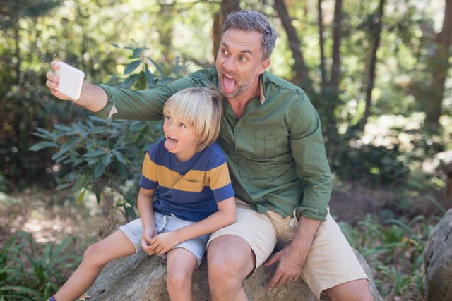 Father and son enjoying a playful moment in the forest, sticking out their tongues while taking a selfie. Ideal for use in family-oriented content, advertisements promoting outdoor activities, or articles about parent-child bonding and fun.