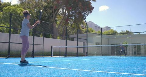 Mature woman swinging paddle on vibrant blue court playing pickleball under clear sky