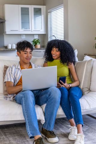 Diverse couple sharing laptop on sofa, remote working and co-browsing at home