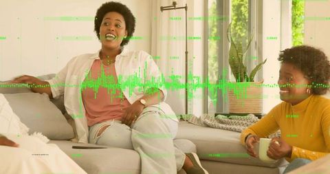 Women laughing and chatting on sunlit sofa with tablet and mug, digital waveform overlay