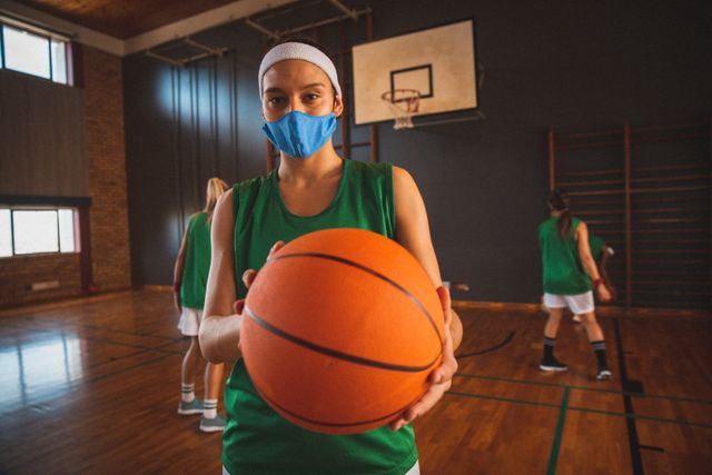Female basketball player wearing a face mask and holding a ball in an indoor court. Other players are seen in the background, also wearing sports uniforms. This image can be used to illustrate sports activities during the COVID-19 pandemic, emphasizing health and safety measures in team sports. Ideal for articles on sports safety, pandemic precautions, and fitness during health crises.