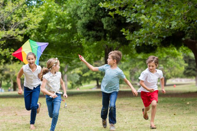 Children running and playing with a colorful kite in a park on a sunny day. Ideal for use in advertisements, educational materials, and websites promoting outdoor activities, family fun, and childhood joy.