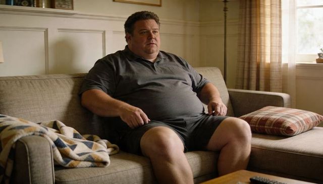 Middle-aged man sitting on beige sofa holding remote wearing dark gray polo and shorts