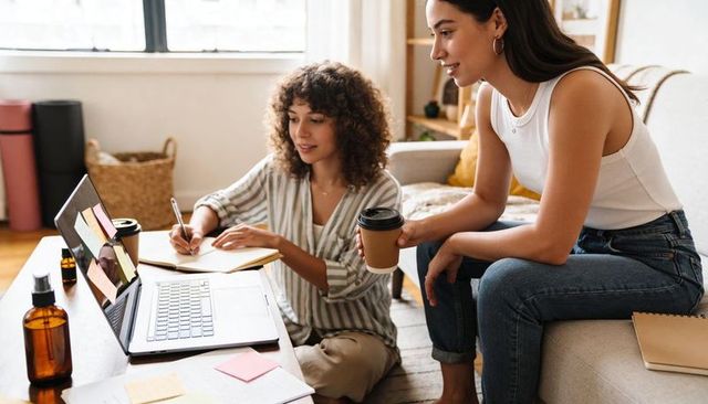 Two women collaborating on laptop and notebook in cozy living room with coffee sharing ideas