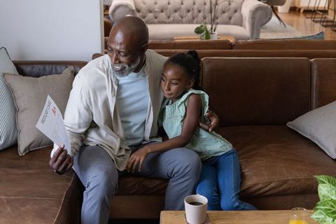 African american grandfather celebrating grade a+ report card while hugging granddaughter on sofa