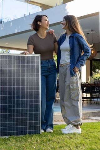 Multiracial women standing with solar panel in backyard celebrating sustainable home energy