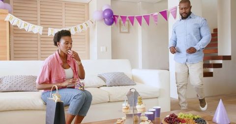 African American couple celebrating birthday at home woman eating candy while partner walking in